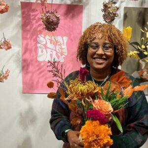 Photo of Cachet smiling among many flowers, alive and dried and a pink poster saying Sun Day